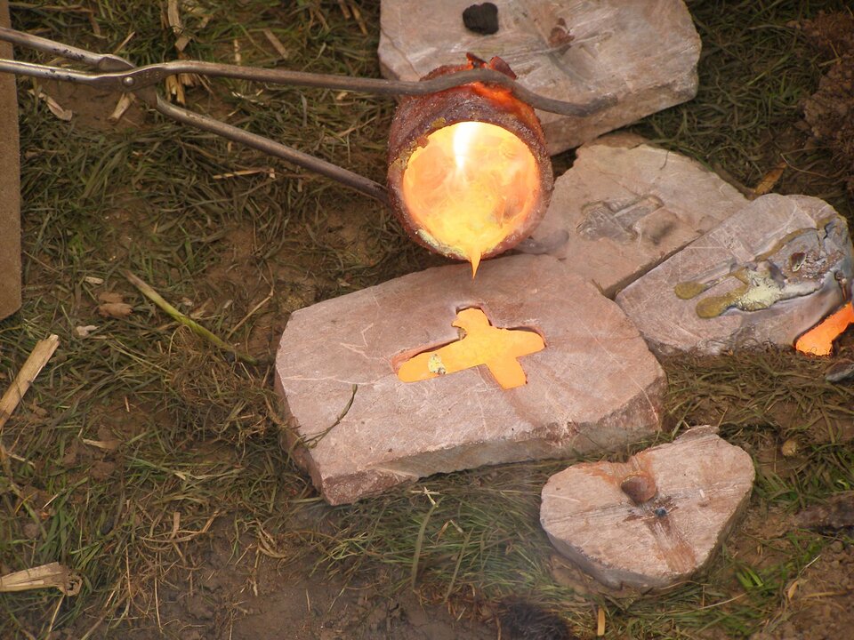 Zdjęcie przedstawia odlewanie przedmiotu z brązu dawnymi metodami. Na obrośniętej trawą ziemi leży wycięta w kamieniu forma w kształcie krzyżyka, do której z trzymanego w długich szczypcach małego tygla nalewany jest płynny metal. Zarówno zawartość formy, jak i tygla jarzą się czerwonawym blaskiem.