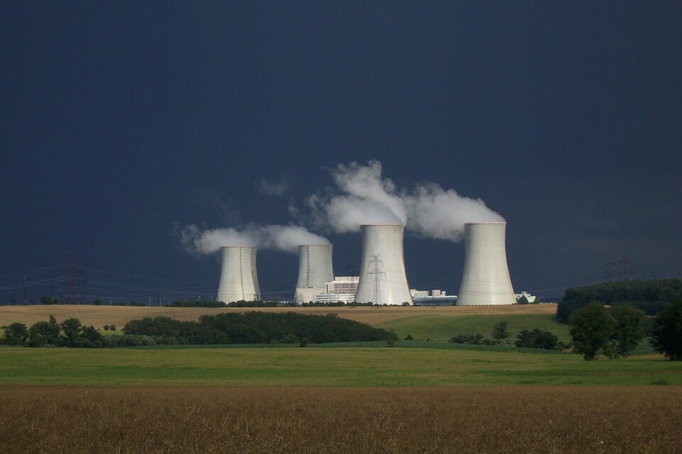 Zdjęcie przedstawia elektrownię atomową widoczną na horyzoncie zdjęcia krajobrazowego. Na pierwszym planie u dołu kadru pola zbóż przetykane niewielkimi lasami. Niebo ciemnoniebieskie. Widoczne cztery kominy chłodnicze, z których wylatują kłęby skroplonej pary wodnej (mgły) i o połowę niższy, duży budynek pomiędzy nimi. Wokół kompleksu elektrowni znajdują się liczne linie energetyczne.