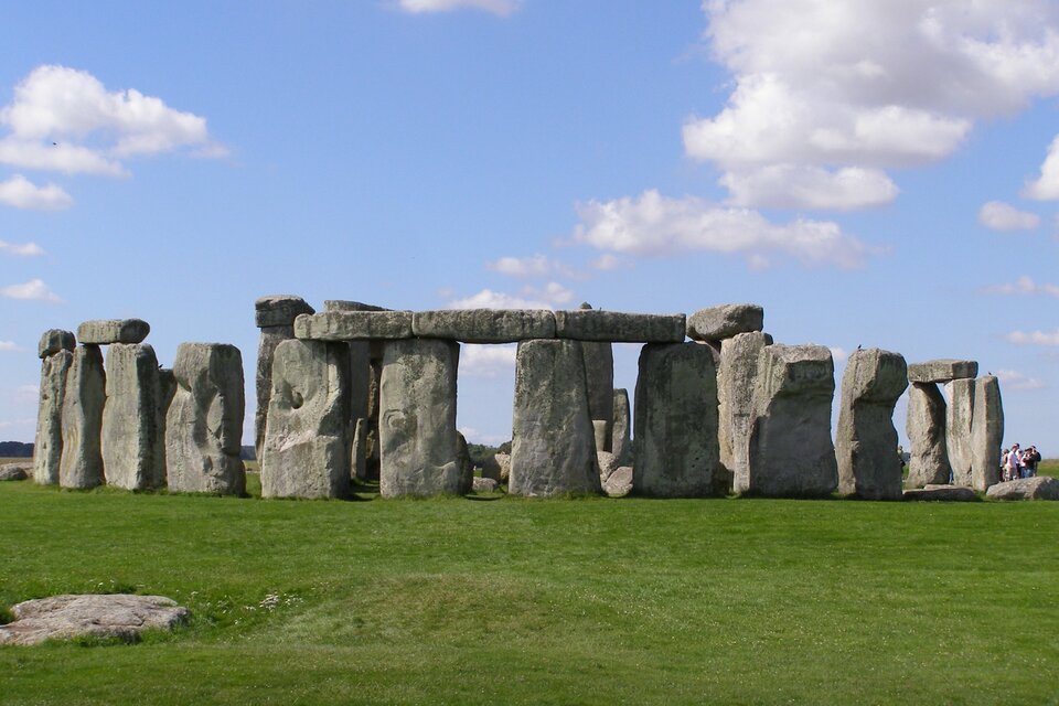 Zdjęcie przedstawia kamienny krąg Stonehenge widziany z odległości kilkudziesięciu metrów. Krąg zajmuje całą szerokość kadru, otaczają go tylko trawa i niebo. Na skraju kadru widoczna jest pomiędzy głazami niewielka grupa ludzi zapewniająca skalę porównawczą dla skał liczących do dziewięciu metrów wysokości.