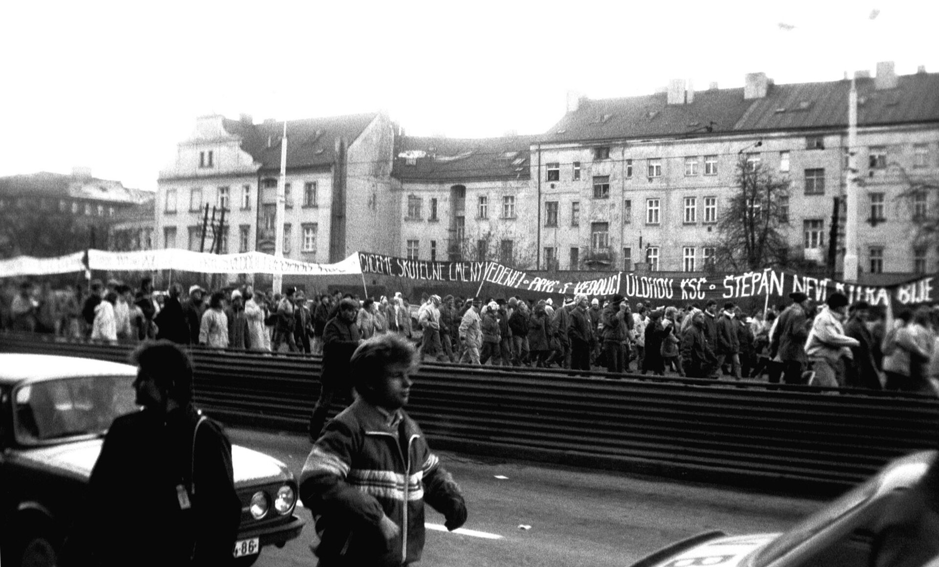 Na czarno-białym zdjęciu demonstracja. Tłum idzie ulicą, transparenty z napisami w języku czeskim.