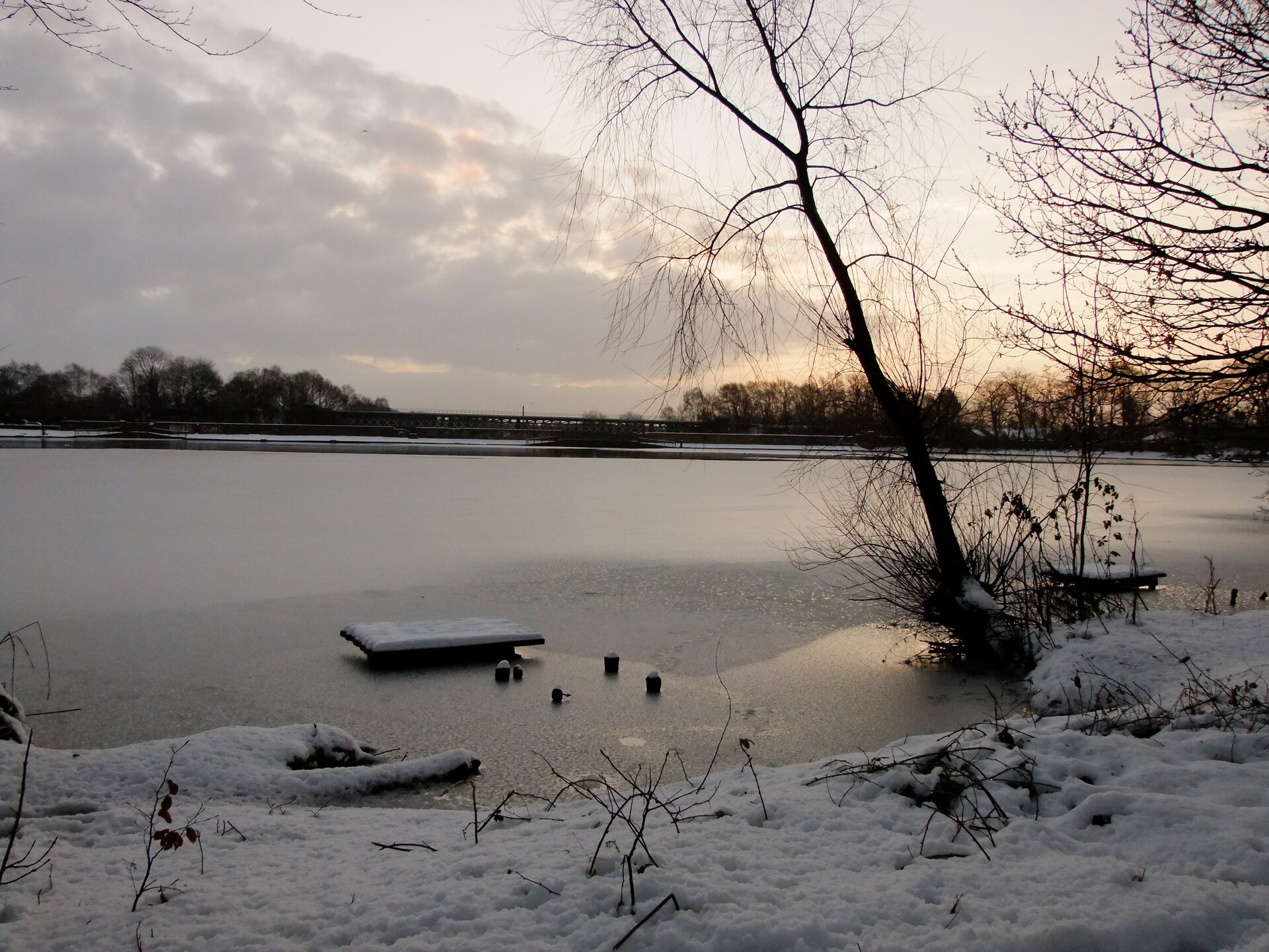 Fotografia wykonana zimą prezentuje duże jezioro oraz fragment jego brzegu okrytego śniegiem. Tafla jeziora jest okryta śniegiem i lodem. Przy samym brzegu nie ma lodu na jeziorze.
