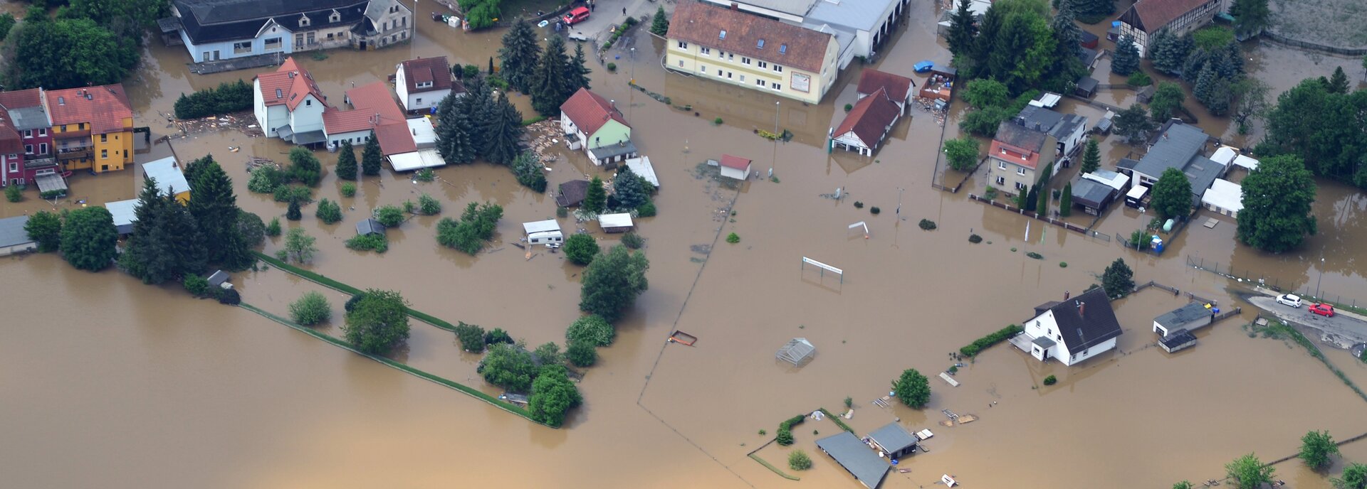 Kolorowe zdjęcie niewielkiej miejscowości zrobione z góry. Cały obszar zalany jest wodą. Z wody, która jest w kolorze brązowym, wystają pojedyncze domy jednorodzinne, budynki, drzewa i samochody.