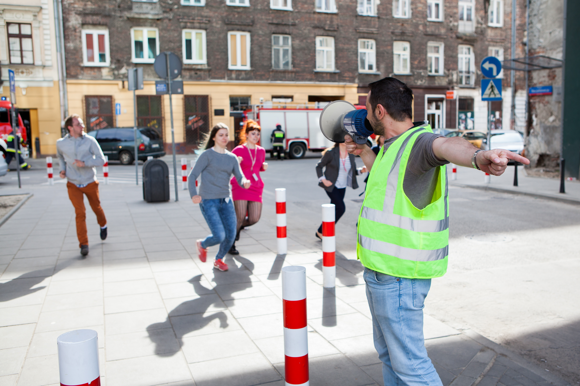 Zdjęcie przedstawiające kierownika ewakuacji wraz z megafonem, który instruuje ludzi gdzie mają się ewakuować. Grupa młodych ludzi biegnie w kierunku wskazywanym przez niego ręką. W tle widać fragment ulicy i pas kamienic oraz stojący na jezdni wóz strażacki.