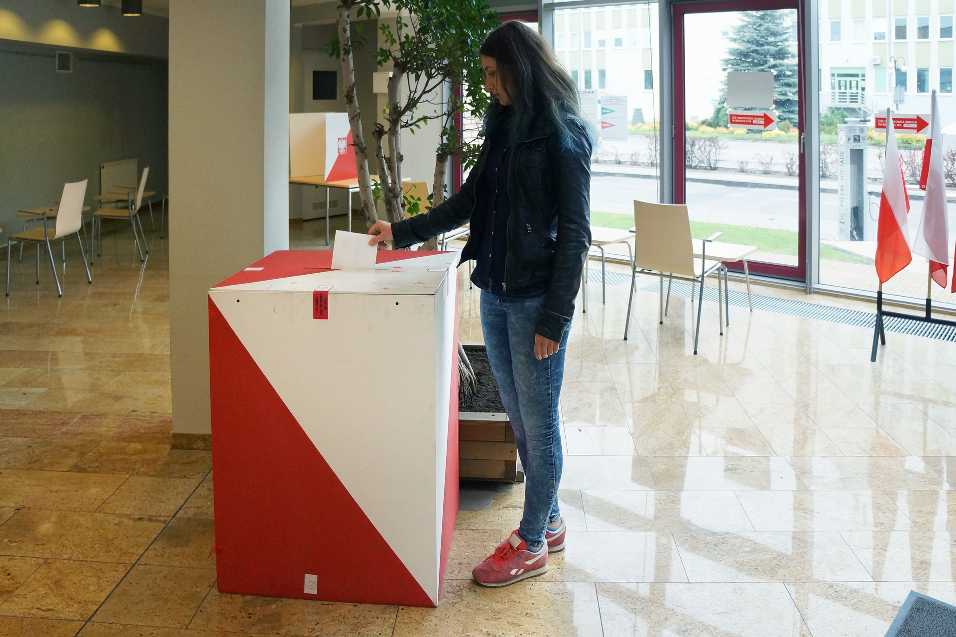 Kolorowe zdjęcie przedstawia dzień wyborów. Duży, dobrze oświetlony hall w budynku. Po prawej stronie okna sięgające od podłogi do sufitu. Na pierwszym planie biało czerwona urna wyborcza. Wysokość urny to około 1 metr. Urna w kształcie prostopadłościanu. W górnej części urny otwór do wrzucania kart z wynikami głosownia wyborców. Na prawo od urny młoda kobieta przodem zwrócona do urny, lewym bokiem do obserwatora zdjęcia. Kobieta wrzuca kartę do głosowania do urny.