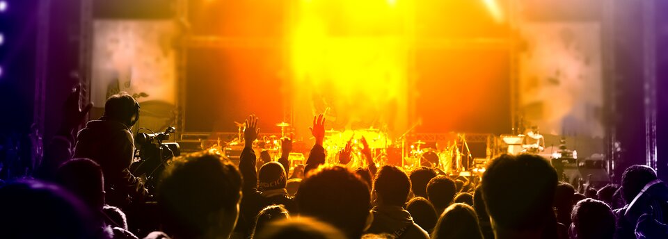 Kolorowe zdjęcie przedstawia scenę i stojący przed nią tłum. Na scenie ustawione są instrumenty muzyczne – daje się odróżnić perkusję, mikrofony. Tłum ludzi zwrócony tyłem do obserwatora, patrzący w kierunku sceny. Widoczne są tylko głowy i wyciągnięte w górę pojedyncze ręce. Po lewej stronie kadru, zwrócony tyłem do obserwatora, siedzi operator kamery, filmujący koncert.