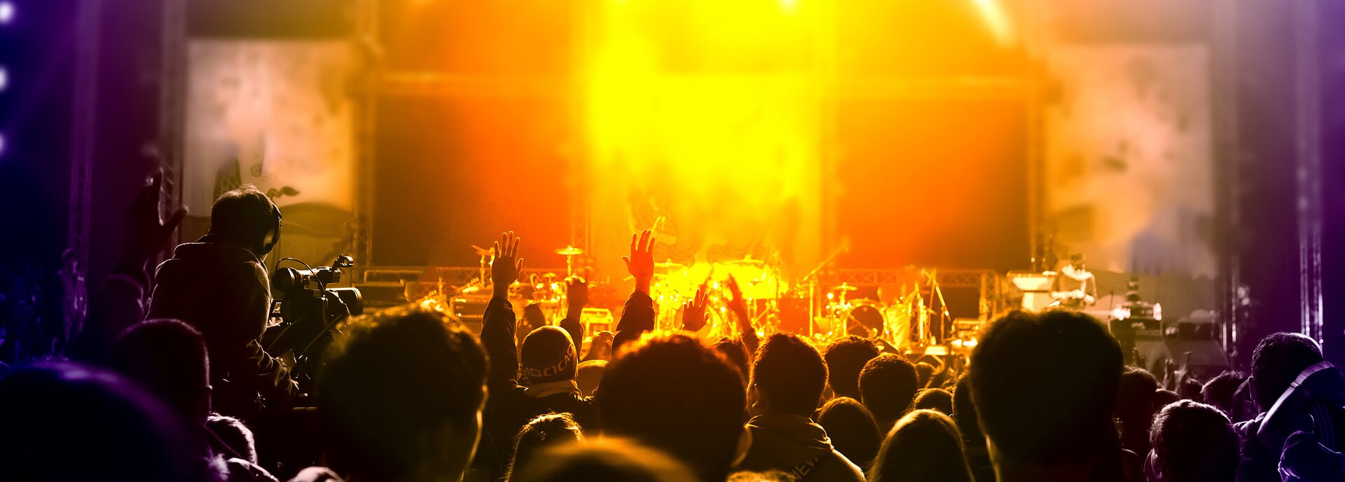 Kolorowe zdjęcie przedstawia scenę i stojący przed nią tłum. Na scenie ustawione są instrumenty muzyczne – daje się odróżnić perkusję, mikrofony. Tłum ludzi zwrócony tyłem do obserwatora, patrzący w kierunku sceny. Widoczne są tylko głowy i wyciągnięte w górę pojedyncze ręce. Po lewej stronie kadru, zwrócony tyłem do obserwatora, siedzi operator kamery, filmujący koncert.