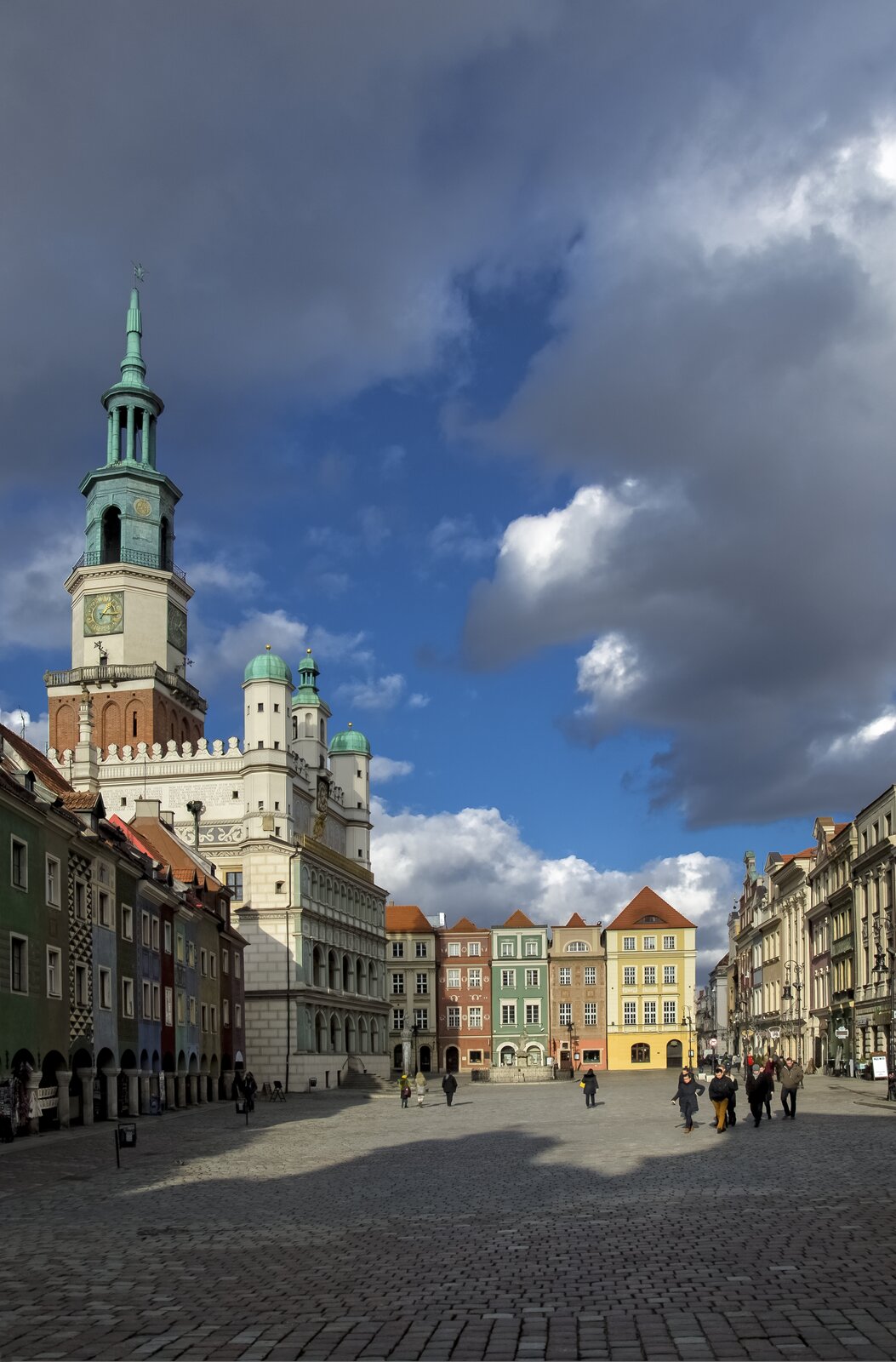 Ilustracja przedstawia ratusz w Poznaniu wraz z zabudową miejską - znajdującymi się w jego pobliżu kamienicami. Po lewej stronie widoczny jest ratusz ze smukłą wieżą zegarową oraz białymi murami, zwieńczonymi w narożach wieżyczkami z hełmami. Po jego lewej stronie znajdują się kamienice z kolorowymi elewacjami. Podobne kamienice usytuowane są przy prostopadłej do ratusza ulicy. Przed ratuszem, po brukowym rynku, spacerują ludzie.