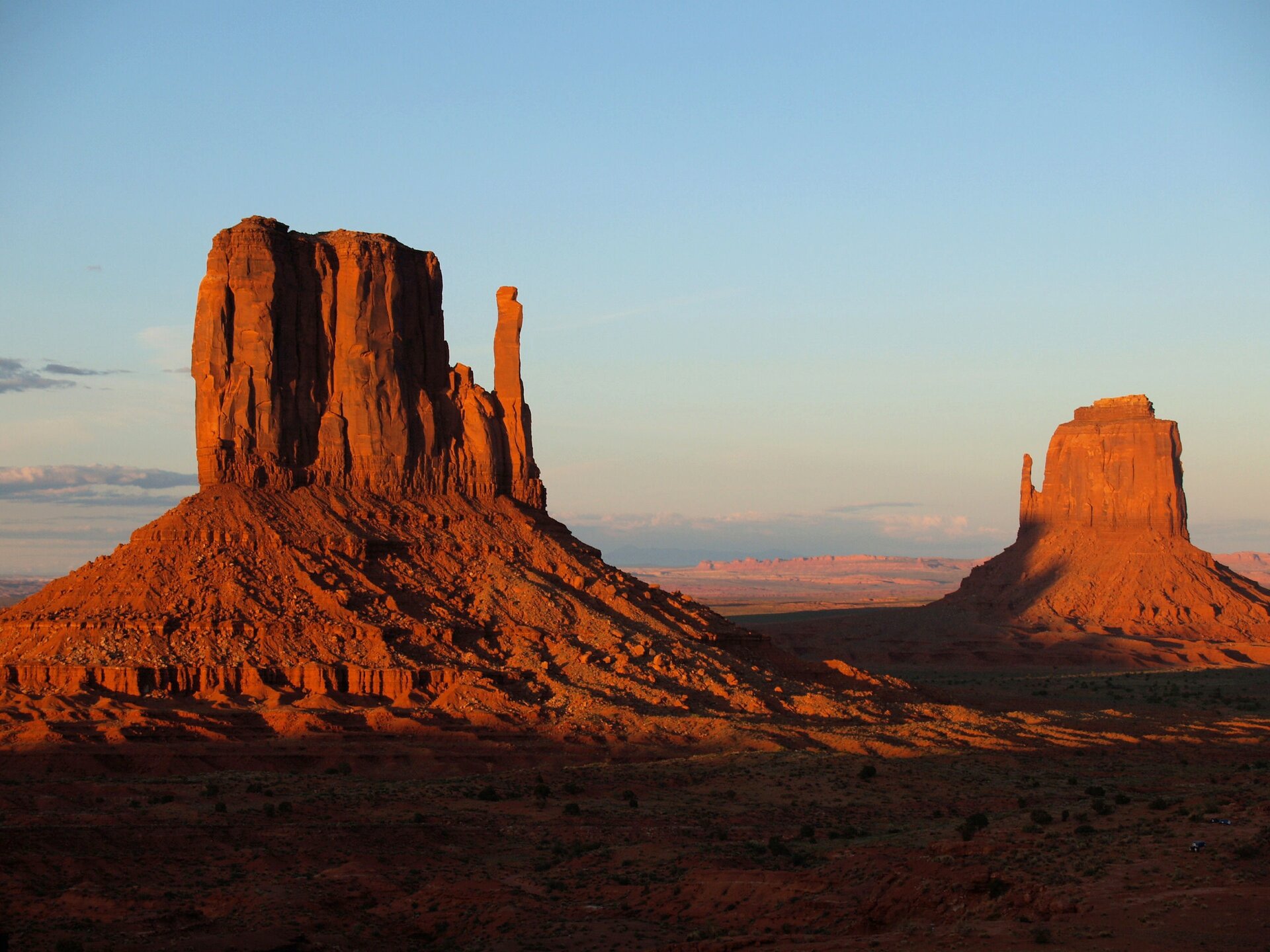 Fotografia prezentuje pustynię skalistą w Monument Valley w USA. Na pierwszym planie widoczne dwie wysokie, prostokątne góry u podstawy, których znajdują się zwietrzałe okruchy skalne. Skały są koloru czerwonego.