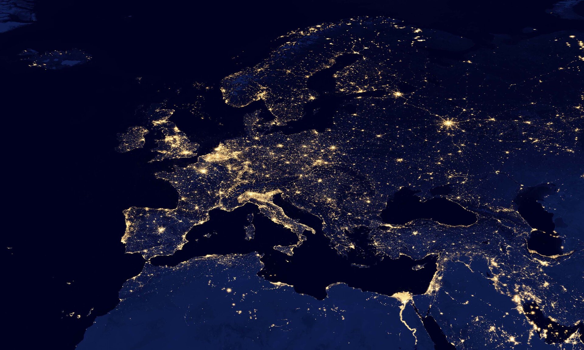 Zdjęcie satelitarne Europy nocą. Na zdjęciu widać zarys kontynentu. Widoczne są światła wielkich aglomeracji. Światła to żółte punkty na tle granatowej Europy. Duże skupiska świateł tworzą żółte plamy. Najbardziej oświetlone są północne Włochy, Niemcy oraz Wyspy Brytyjskie. Pojedyncze punkty to mniejsze miasta.