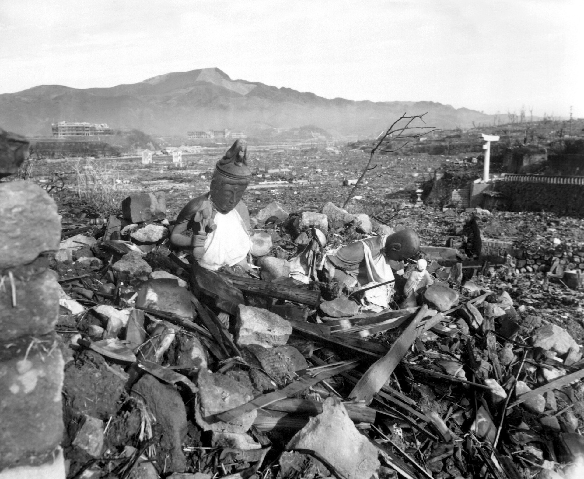 Zdjęcie przedstawia zniszczone przez wybuch bomby atomowej tereny Hiroszimy i Nagasaki. Fotografia czarno-biała. Na pierwszym planie widoczne starta gruzów, połamanych desek i konstrukcji oraz fragment figurki Buddy. Nieco dalej widać ogrom zniszczeń spowodowanych przez wybuch wojny. Prawie wszystko zostało zrównane z ziemią. W oddali widać szkielety budynków. W tle zarys gór.