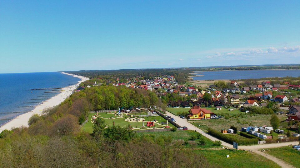 Na zdjęciu widok z góry na brzeg morski, plażę, teren zabudowany i tereny zielone oraz jezioro przybrzeżne w niewielkiej odległości od morza.