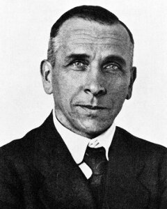 Na zdjęciu Alfred Wegener. Mężczyzna w średnim wieku. Sylwetka od ramion w górę. Ciemne, krótkie włosy, zaczesane do tyłu. Ubrany w garnitur, białą koszulę i krawat.