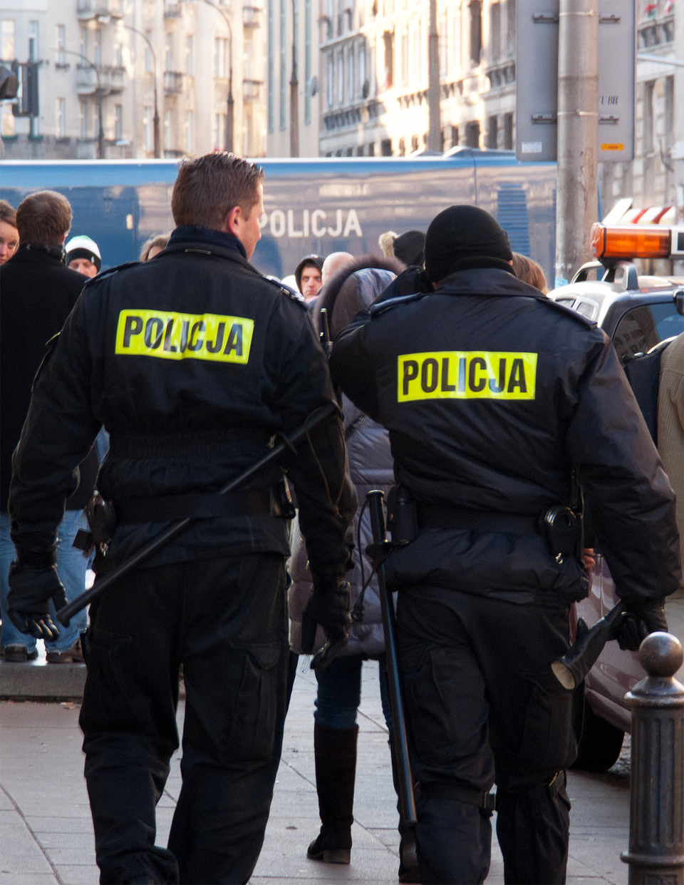 Kolorowe zdjęcie przedstawia dwóch policjantów tyłem do obserwatora zdjęcia. Słoneczny dzień. Policjanci idą wzdłuż chodnika w centrum miasta. Ubrani w czarne mundury. Na plecach żółty odblaskowy prostokąt z czarnym napisem policja. W głębi tłum ludzi oraz wysokie budynki w mieście. Po prawej stronie dach wozu policyjnego ze światłami ostrzegawczymi na dachu wozu.