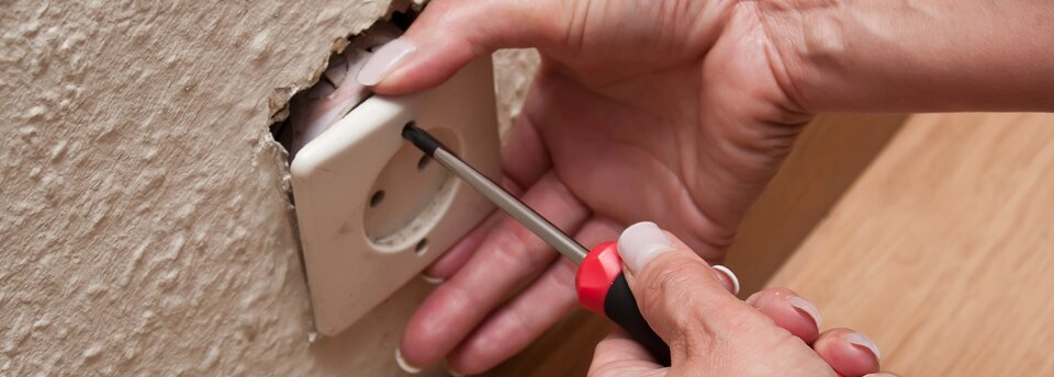 Kolorowe zdjęcie przedstawia wyjęte ze ściany gniazdko elektryczne podtrzymywane przez jedną dłoń. Osoba podtrzymująca gniazdko ma w drugiej dłoni śrubokręt włożony w otwór, którym można gniazdko przymocować do ściany.