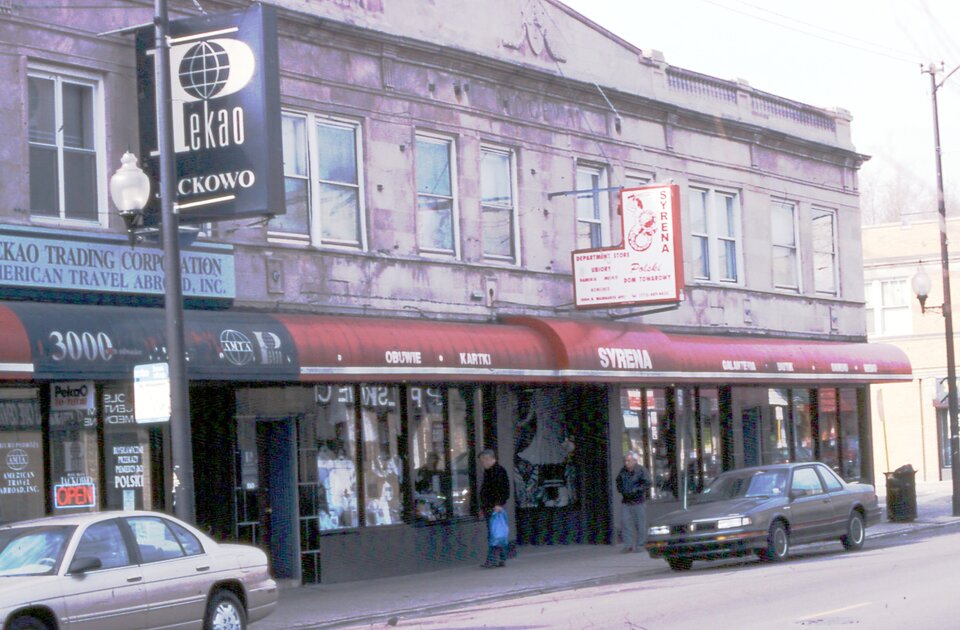 Na zdjęciu ulica w Chicago. Nowoczesne budynki mieszkalno-usługowe, na szyldach napisy w języku polskim. Na ulicach samochody, kilku przechodniów.