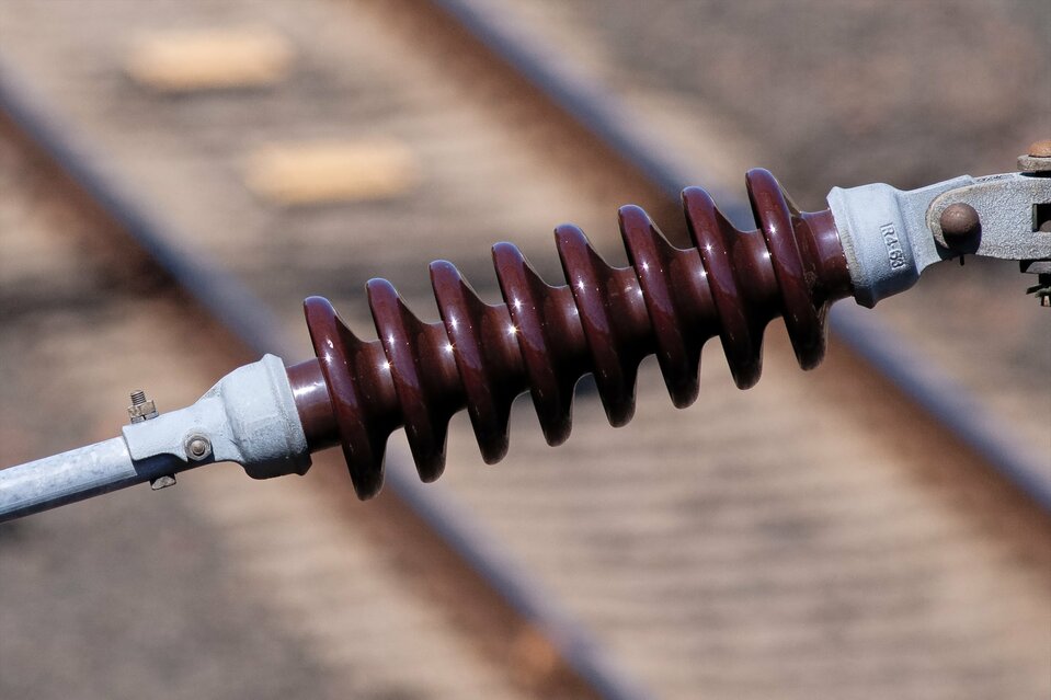 Zdjęcie przedstawia w zbliżeniu ceramiczny izolator łączący dwa elementy metalowe kolejowej sieci trakcyjnej. W tle w obszarze nieostrym zdjęcia można rozpoznać tory kolejowe. Sam izolator ma kolor ciemnoczerwony, a kształtem przypomina ceramiczny pręt z licznymi następującymi po sobie zgrubieniami o kształtach kapeluszy grzybów. Zgrubienia te mają za zadanie zapobiegać przenoszeniu się przez powietrze wyładowań w postaci iskier.