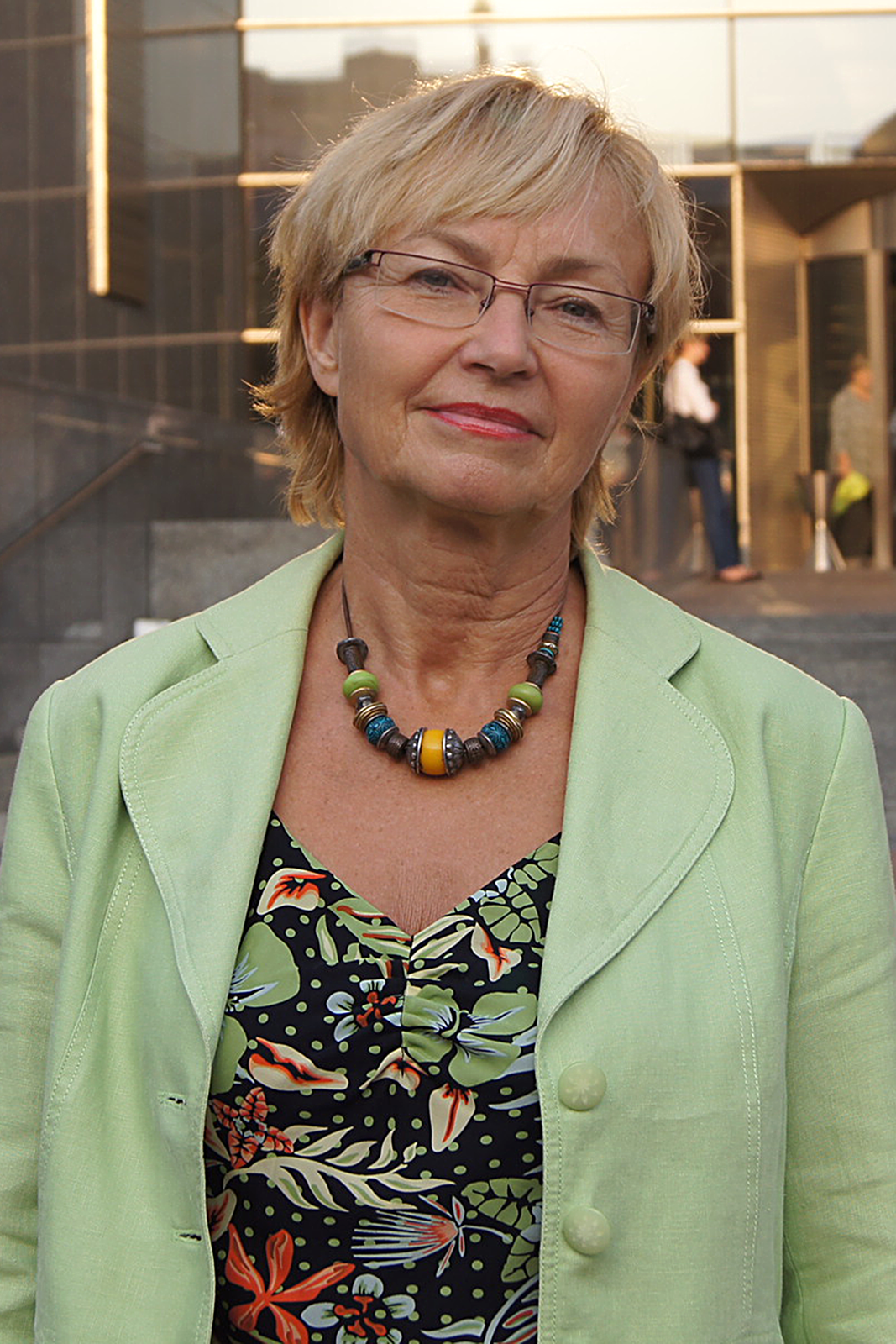 Kolorowe zdjęcie portretowe Leny Barbary Kolarskiej-Bobińskiej. Kobieta w wieku ponad 60 lat, przodem do obserwatora zdjęcia. Włosy proste blond z lekko opadającą grzywką na czoło. Krótkie włosy zaczesane za uszy. Czoło wysokie. Brwi ciemne. Nos duży. Usta wąskie. Okulary w metalowych oprawkach. Twarz okrągła. Ubrana w kwiecistą bluzkę i jasno zieloną marynarkę. Na szyi wisiorek z drewnianych niebieskich i zielonych korali.