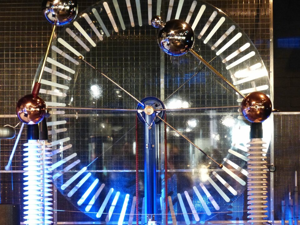 Zdjęcie przedstawia maszynę elektrostatyczną, której głównym elementem konstrukcyjnym są dwie sąsiadujące ze sobą obracające się tarcze. Na zdjęciu wykonane są one z przezroczystego materiału, dzięki czemu dobrze widoczne są jedynie koncentrycznie ułożone wzdłuż krawędzi aluminiowe paski, które podczas obrotu tarcz w przeciwnych kierunkach ulegają elektryzacji. Przed tarczami na ruchomych ramionach znajdują się metalowe kulki iskiernika, które można zbliżać lub oddalać od siebie.