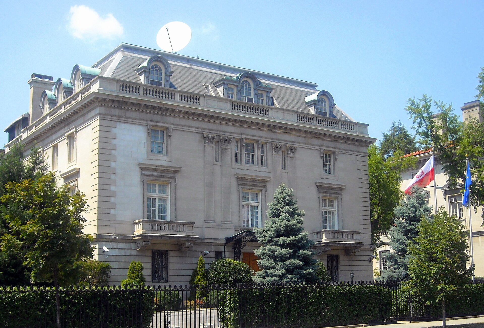 Kolorowe zdjęcie przedstawia piętrowy budynek ambasady Polski w Waszyngtonie. Budynek z białego kamienia. Na pierwszym piętrze duże okna. Przed oknami balustrady balkonów. Na pierwszym piętrze małe okna ozdobione rzeźbami powyżej. Wokół dachu balustrada z kamienia. W dachu okna w formie wykuszy. Okna zaokrąglone w górnej części. Dach pokryty szarą dachówką. Na dachu duża antena satelitarna. Przed budynkiem drzewa i krzewy. Budynek otoczony czarnym kutym płotem. Wzdłuż płotu, od podwórza, zielony żywopłot. Po prawej stronie, przed budynkiem, dwa maszty z flagami. Po lewej flaga Polski. Na fladze godło Polski. Po prawej flaga Unii Europejskiej.