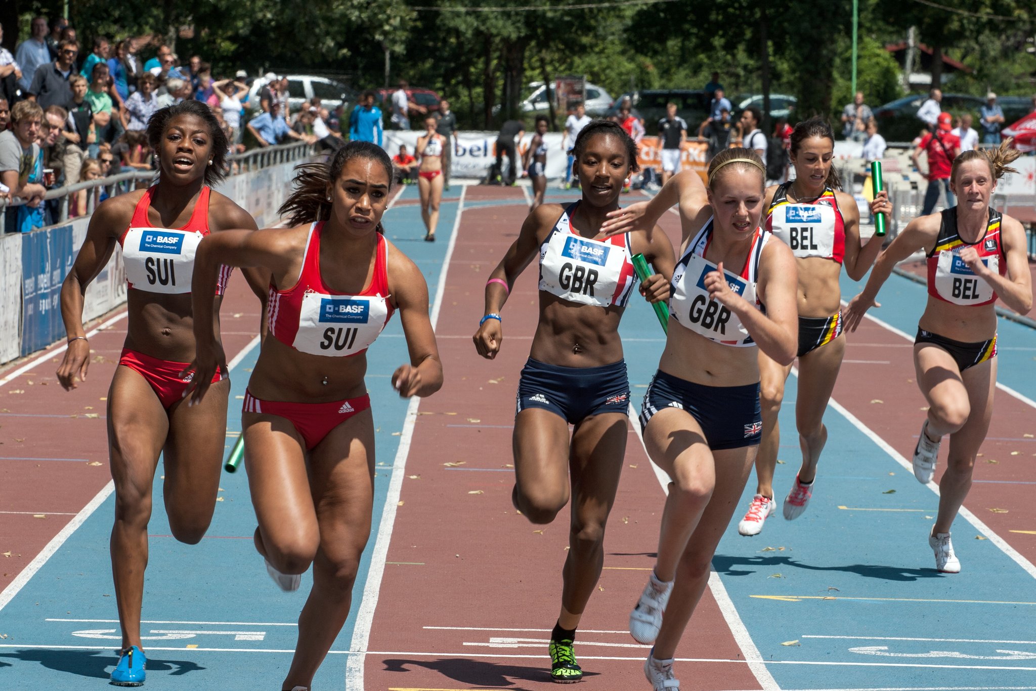 Na bieżni lekkoatletycznej w miejscu startu znajdują się biegaczki trzech drużyn. Dwie mają na piersiach napis SUI, dwie GBR a dwie BEL. Biegaczki biegną w sztafecie i pokazany jest moment, w którym biegaczki wcześniejszej zmiany przekazują pałeczkę biegaczkom kolejnej zmiany. W oddali widać inne biegaczki na bieżni, a po lewej stronie za barierkami stoją widzowie.