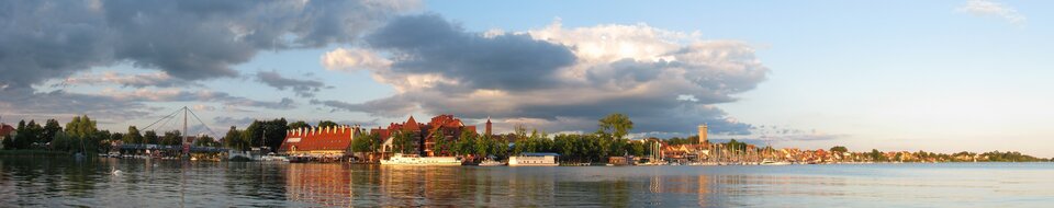 Na zdjęciu panorama miasta nad brzegiem zbiornika wodnego. W tle zabudowania, wieże, przy brzegu zacumowane jachty i łódki.